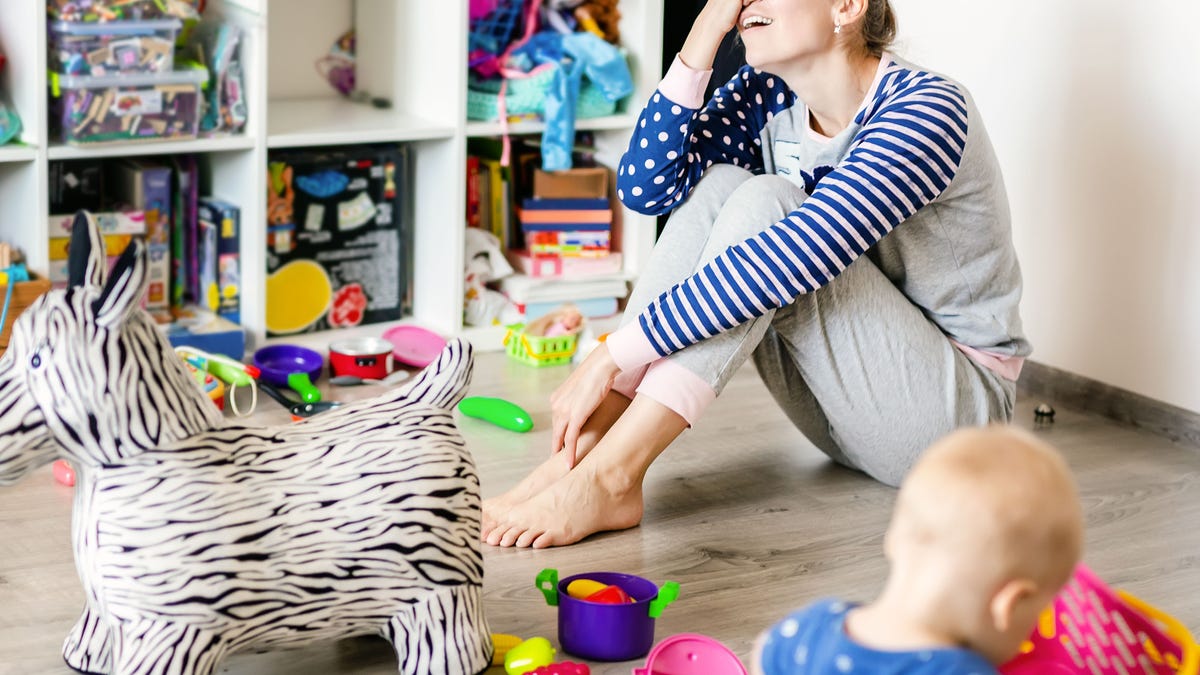 Tired of everyday household mother sitting on floor with hands on face. Kid playing in messy room. Scaterred toys and disorder. Happy parenting