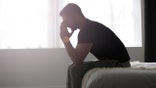 A man sitting at the end of a bed, deep in solemn thought.