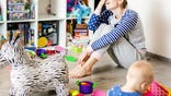 Tired of everyday household mother sitting on floor with hands on face. Kid playing in messy room. Scaterred toys and disorder. Happy parenting