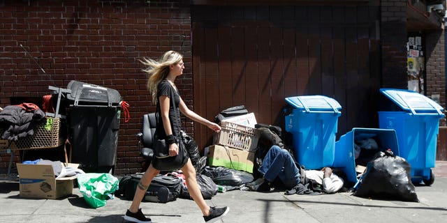 A woman walks past a homeless man sleeping in front of recycling bins and garbage on a street corner in San Francisco. 