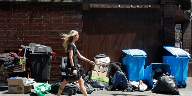 A woman walks past a homeless man sleeping in front of recycling bins and garbage on a street corner in San Francisco.