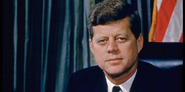 Le président John F. Kennedy en posant pour une photo à son bureau avec un drapeau américain en arrière-plan. 
