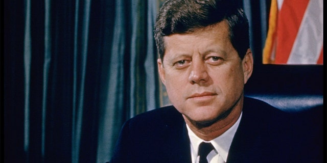 President John F. Kennedy while posing for a picture at his desk with a U.S. flag in the background.