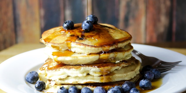 A stack of homemade blueberry pancakes with fresh blueberries and maple syrup is shown here. Busy moms and dads can defrost and then reheat pancakes that are made ahead of time.