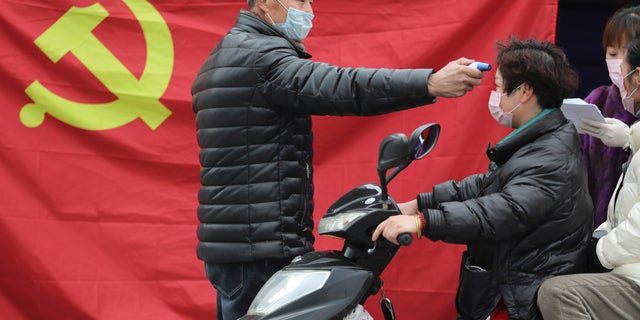 A volunteer stands in front of a Communist Party flag as he takes the temperature of a scooter driver at a roadside checkpoint in Hangzhou in eastern China's Zhejiang Province, Monday, Feb. 3, 2020. China sent medical workers and equipment to a newly built hospital, infused cash into financial markets and further restricted people's movement in sweeping new steps Monday to contain a rapidly spreading virus and its escalating impact. (Chinatopix via AP)