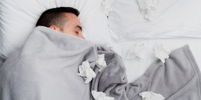Ill young man in bed, covered with a light gray blanket, surrounded by used tissues