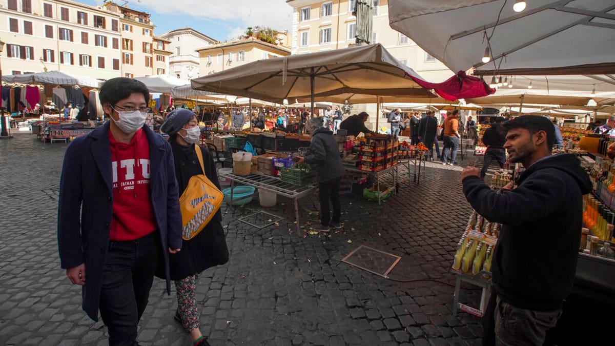A seller calls for customers as a couple wearing face masks walks by, at the Campo de' Fiori street market, in Rome, Saturday, March 7, 2020. 
