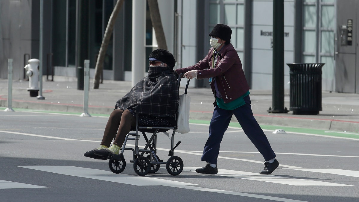 A woman wearing a mask pushes a person in a wheelchair across a street in San Francisco. (AP Photo/Jeff Chiu)