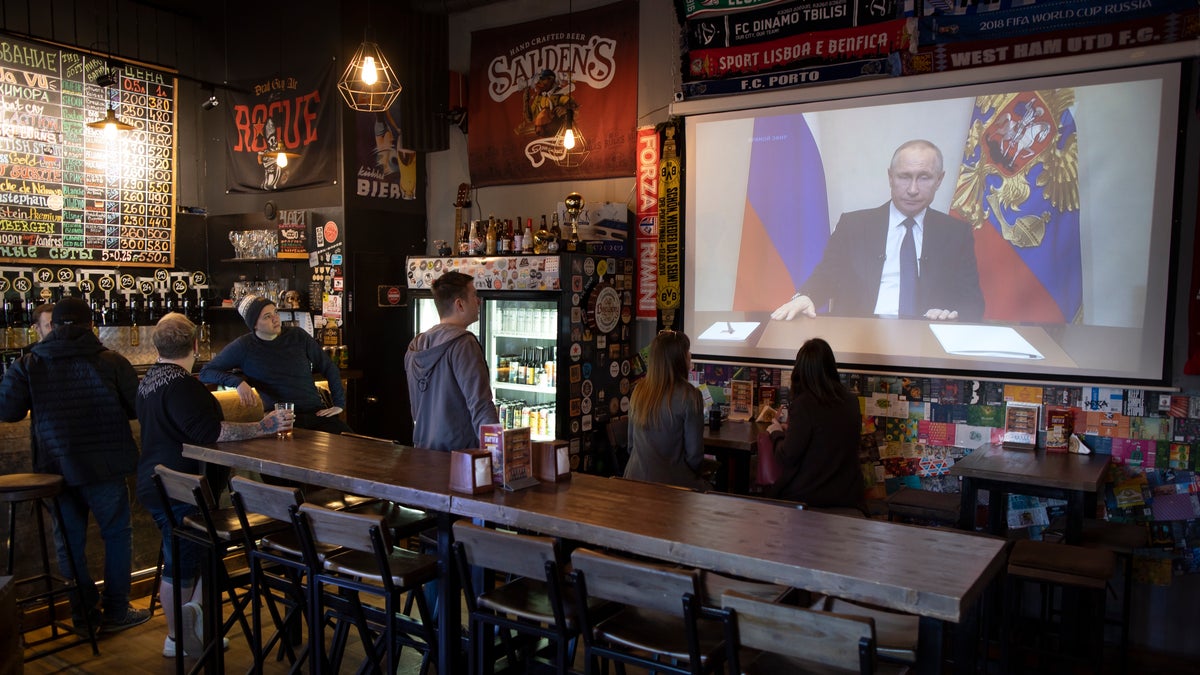 A few visitors and staff of the pub watching the broadcast of Russian President Vladimir Putin addresses Russian citizens on the State Television channels in Moscow, Russia, Wednesday, March 25, 2020. Putin has postponed a nationwide vote on proposed constitutional amendments that include a change that would allow him to seek another term in power. He also announced during a televised address to the nation that the government doesn't want Russians except those working in essential sectors to come to work next week. He said that stores, pharmacies and banks will stay open.