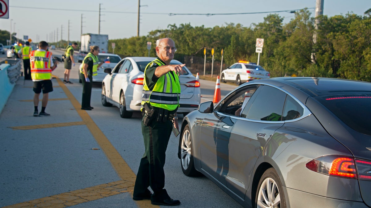 In this photo provided by the Florida Keys News Bureau, Monroe County Sheriff’s Office Col. Lou Caputo directs a driver wanting to continue down the Florida Keys Overseas Highway near Key Largo, Fla. Friday, March 27, 2020. The Keys have been temporarily closed to visitors since March 22, because of the coronavirus crisis. Keys officials decided to established the checkpoint Friday to further lessen the threat of virus transmission to people in the subtropical island chain.