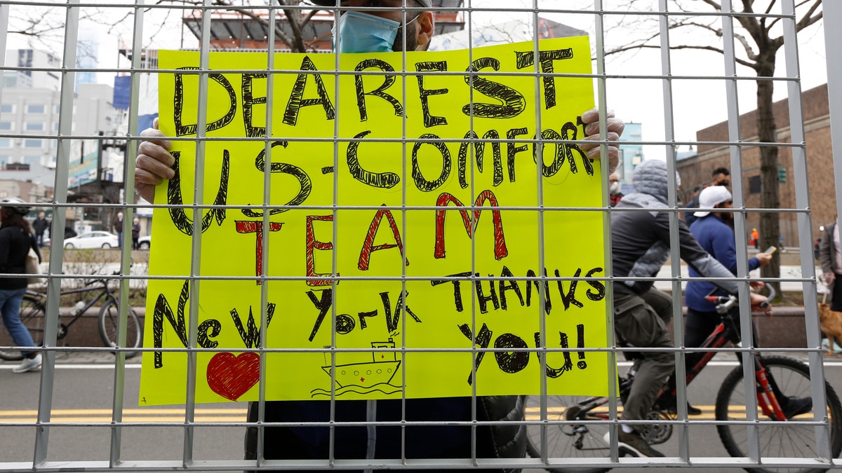 Irakli Kalarji holds a sign against a fence at Pier 90 during the arrival of the USNS Comfort, a naval hospital ship with a 1,000 bed-capacity, Monday, March 30, 2020, in New York. (AP Photo/Kathy Willens)