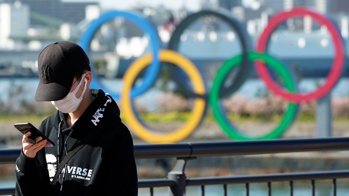 In this March 3, 2020, photo, a tourist wearing a protective mask takes a photo with the Olympic rings in the background, at Tokyo's Odaiba district in Tokyo.  (AP Photo/Eugene Hoshiko)