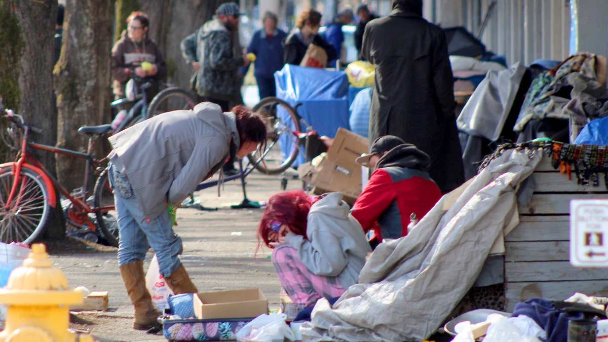 In this Tuesday, March 3, 2020 photo, homeless people crowd a sidewalk in downtown Salem, Ore., where they have set up a makeshift camp. Experts say that the homeless, who often have health and substance-abuse problems, are exposed to the elements and do not have easy access to hygiene, are more vulnerable to the coronavirus. Some cities are making provisions so the homeless who contract the virus have a place to recover without spreading the infection further. (AP Photo/Andrew Selsky)