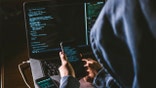 anonymous person in the hood sitting in front of computer working with laptop and mobile phone
