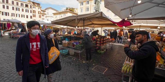 A seller calls for customers as a couple wearing face masks walks by, at the Campo de' Fiori street market, in Rome, Saturday, March 7, 2020. 