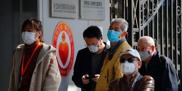 People wearing protective face masks wait in a queue to get temperature check before entering a bank in Beijing, Wednesday, March 11, 2020. For most, the coronavirus causes only mild or moderate symptoms, such as fever and cough. But for a few, especially older adults and people with existing health problems, it can cause more severe illnesses, including pneumonia. 