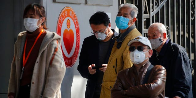 People wearing protective face masks wait in a queue to get temperature check before entering a bank in Beijing, Wednesday, March 11, 2020. For most, the coronavirus causes only mild or moderate symptoms, such as fever and cough. But for a few, especially older adults and people with existing health problems, it can cause more severe illnesses, including pneumonia. 