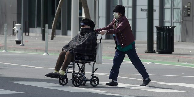 A woman wearing a mask pushes a person in a wheelchair across a street in San Francisco. (AP Photo/Jeff Chiu)