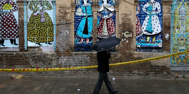 A man shelters from the rain with an umbrella as he walks past an old building decorated with a replica of Iranian old paintings in a mostly empty street in a commercial district in downtown Tehran, Iran, Sunday, March 22, 2020. On Sunday, Iran imposed a two-week closure on major shopping malls and centers across the country to prevent spreading the new coronavirus. Pharmacies, supermarkets, groceries and bakeries will remain open. (AP Photo/Vahid Salemi)