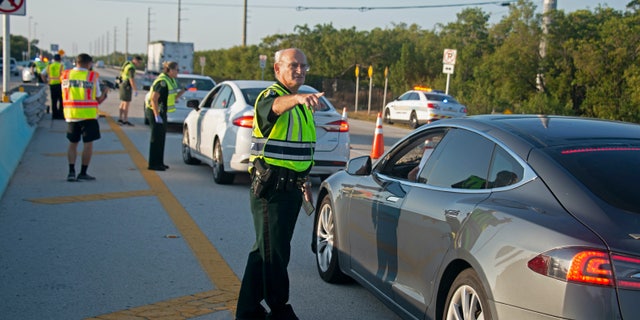 In this photo provided by the Florida Keys News Bureau, Monroe County Sheriff’s Office Col. Lou Caputo directs a driver wanting to continue down the Florida Keys Overseas Highway near Key Largo, Fla. Friday, March 27, 2020. The Keys have been temporarily closed to visitors since March 22, because of the coronavirus crisis. Keys officials decided to established the checkpoint Friday to further lessen the threat of virus transmission to people in the subtropical island chain.