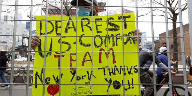 Irakli Kalarji holds a sign against a fence at Pier 90 during the arrival of the USNS Comfort, a naval hospital ship with a 1,000 bed-capacity, Monday, March 30, 2020, in New York. (AP Photo/Kathy Willens)