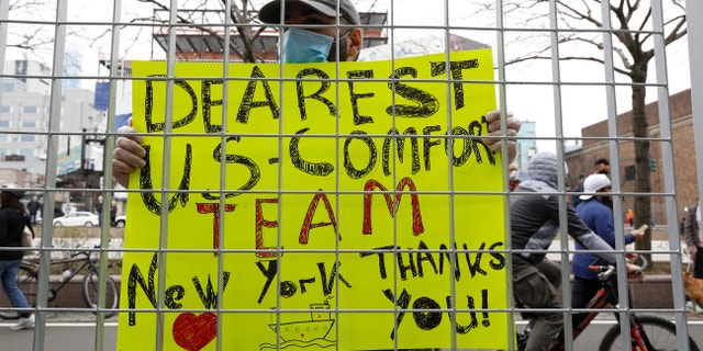 Irakli Kalarji holds a sign against a fence at Pier 90 during the arrival of the USNS Comfort, a naval hospital ship with a 1,000 bed-capacity, Monday, March 30, 2020, in New York. (AP Photo/Kathy Willens)