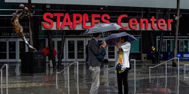 Tourist stand under umbrellas in rain outside Staples Center, home to hockey's Los Angeles Kings and basketball's Los Angeles Lakers, Clippers and Sparks, in Los Angeles on Thursday, March 12, 2020. Officials have banned large gatherings and events in Los Angeles to try to stop the spread of the new coronavirus. (AP Photo/Damian Dovarganes)