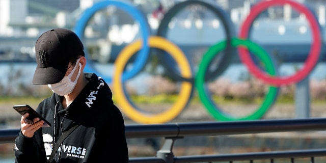 In this March 3, 2020, photo, a tourist wearing a protective mask takes a photo with the Olympic rings in the background, at Tokyo's Odaiba district in Tokyo. (AP Photo/Eugene Hoshiko)