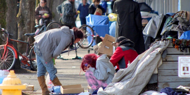 In this Tuesday, March 3, 2020 photo, homeless people crowd a sidewalk in downtown Salem, Ore., where they have set up a makeshift camp. Experts say that the homeless, who often have health and substance-abuse problems, are exposed to the elements and do not have easy access to hygiene, are more vulnerable to the coronavirus. Some cities are making provisions so the homeless who contract the virus have a place to recover without spreading the infection further. (AP Photo/Andrew Selsky)