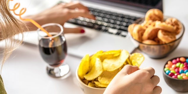 A young woman works at a computer and eats an array of unhealthy foods. Finding the true trigger of a headache is complicated because other factors need to be considered. 