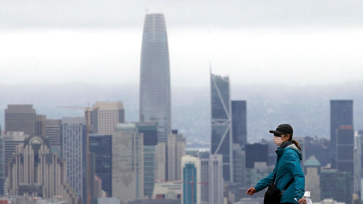 A woman wears a mask during the coronavirus outbreak while crossing a street in front of the skyline in San Francisco, Saturday, April 4, 2020. (AP Photo/Jeff Chiu)