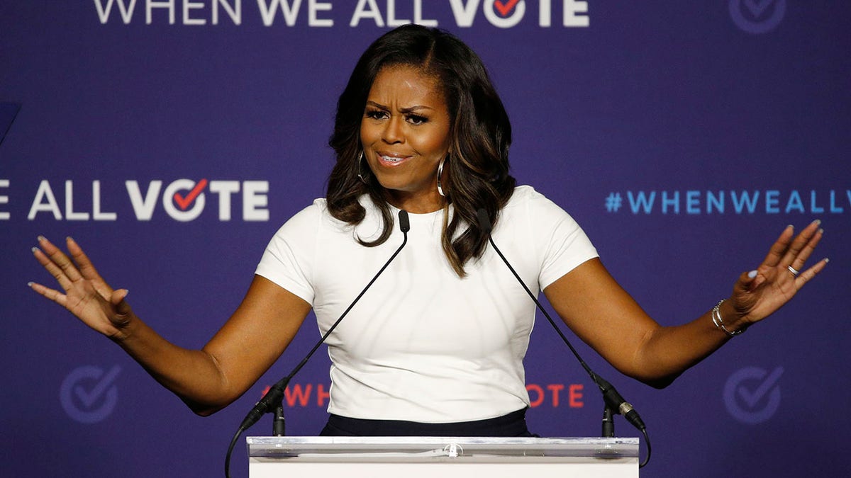 Michelle Obama stands at a podium wearing a white top in front of a "When we all vote" purple background
