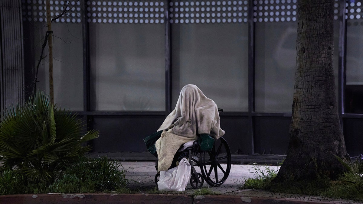 In this April 6, 2020, file photo, a homeless person sits on a wheelchair in rainy weather on Sunset Boulevard in Los Angeles.