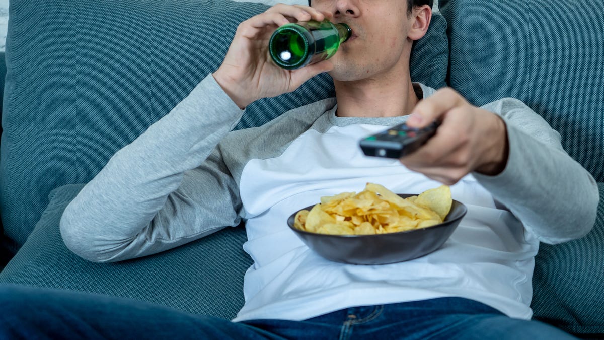 Lifestyle portrait of young bored man on couch with remote control zapping for movie or live sport. Looking disinterested drinking beer. Sedentary and mass social media or Television addiction.