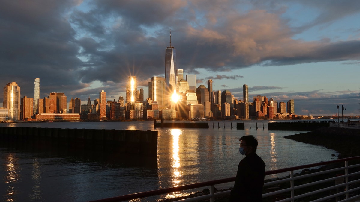 A person in a medical mask watches the sun set on the skyline of lower Manhattan and One World Trade Center in New York City on April 13, 2020 as seen from Jersey City, N.J.