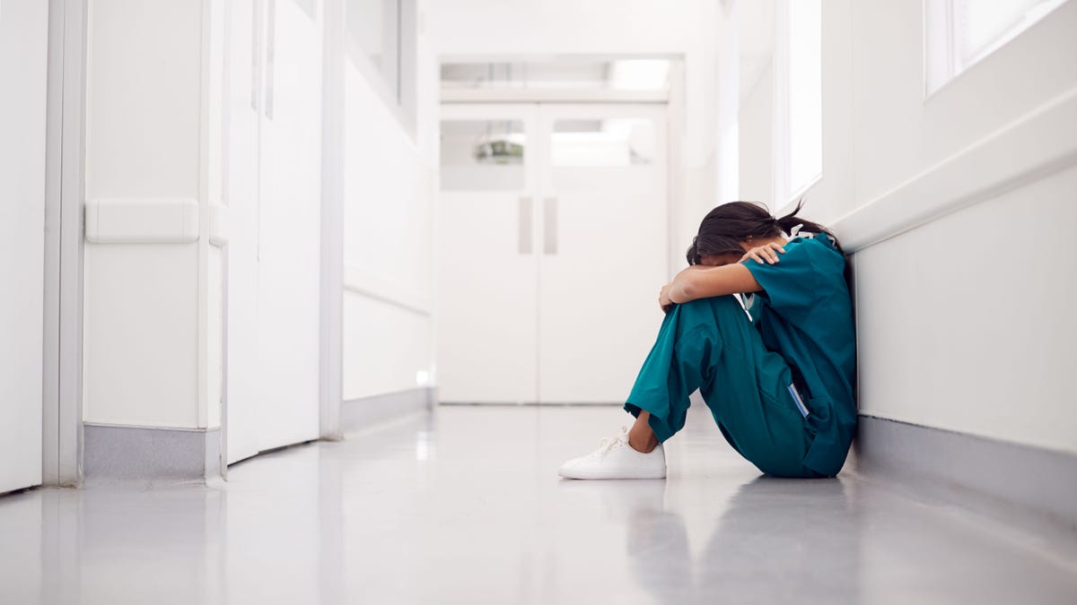 Stressed And Overworked Female Doctor Wearing Scrubs Sitting On Floor In Hospital Corridor