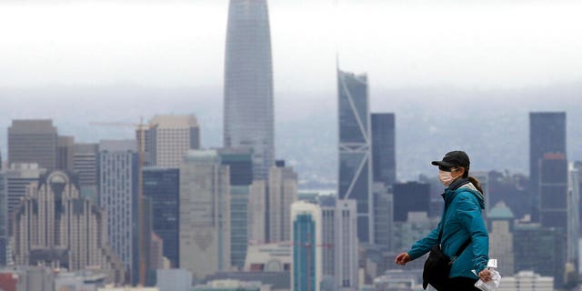 A woman wears a mask during the coronavirus outbreak while crossing a street in front of the skyline in San Francisco, Saturday, April 4, 2020. (AP Photo/Jeff Chiu)