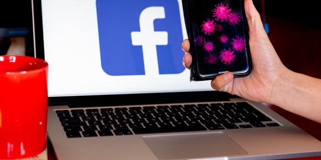 A woman holds her mobile phone in front of a computer screen with facebook logo for illustration purposes during Coronavirus pandemic, Ostrowiec Swietokrzyski on April 14, 2020.