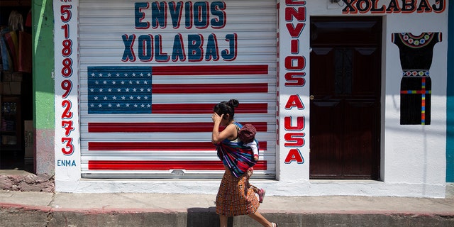 In this April 3, 2020 photo, a woman carrying a child walks past a closed courier business featuring a U.S. flag and the Spanish phrase: "Send to U.S.A" in the largely indigenous town of Joyabaj, Guatemala, where half of the residents depend on remittances, almost all from the U.S.  (AP Photo/Moises Castillo)