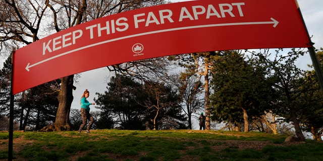 A woman jogs in Brooklyn's Fort Greene Park, Sunday, April 5, 2020, beneath a sign demonstrating the distance people should keep from each other during the coronavirus outbreak in New York.