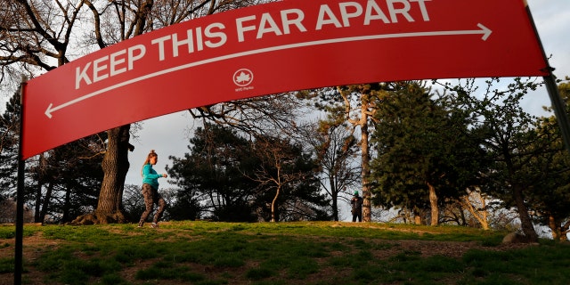 A woman jogs in Brooklyn's Fort Greene Park, Sunday, April 5, 2020, beneath a sign demonstrating the distance people should keep from each other during the coronavirus outbreak in New York.