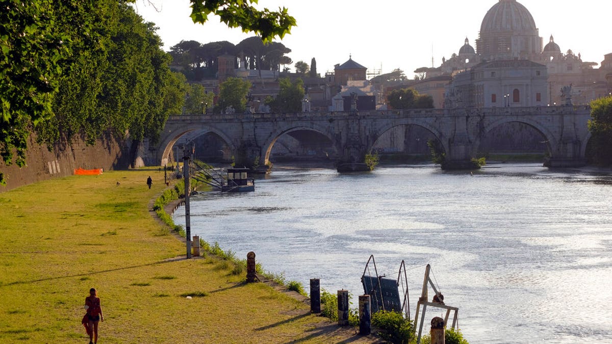 A woman walks along the banks of Tiber river as the dome of St. Peter's Basilica stands out at background right, in Rome, Wednesday, May 6, 2020.