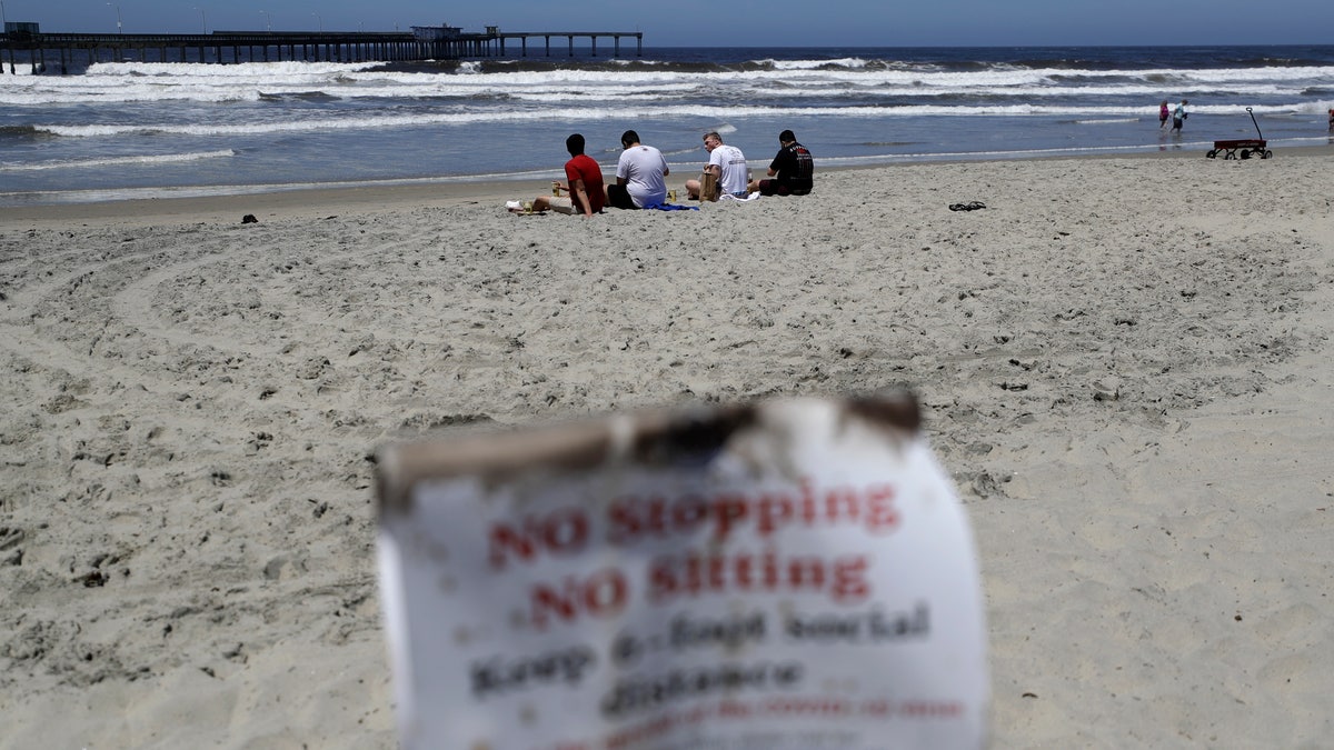 Four men sit behind a sign that reads, "no stopping, no sitting," on a beach closed to stopping, sitting or lying down Wednesday, May 6, 2020, in San Diego. (AP Photo/Gregory Bull)