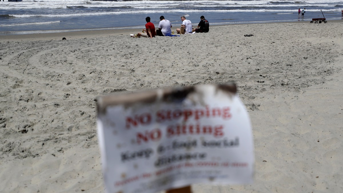 Four men sit behind a sign that reads, "no stopping, no sitting," on a beach closed to stopping, sitting or lying down Wednesday, May 6, 2020, in San Diego. (AP Photo/Gregory Bull)