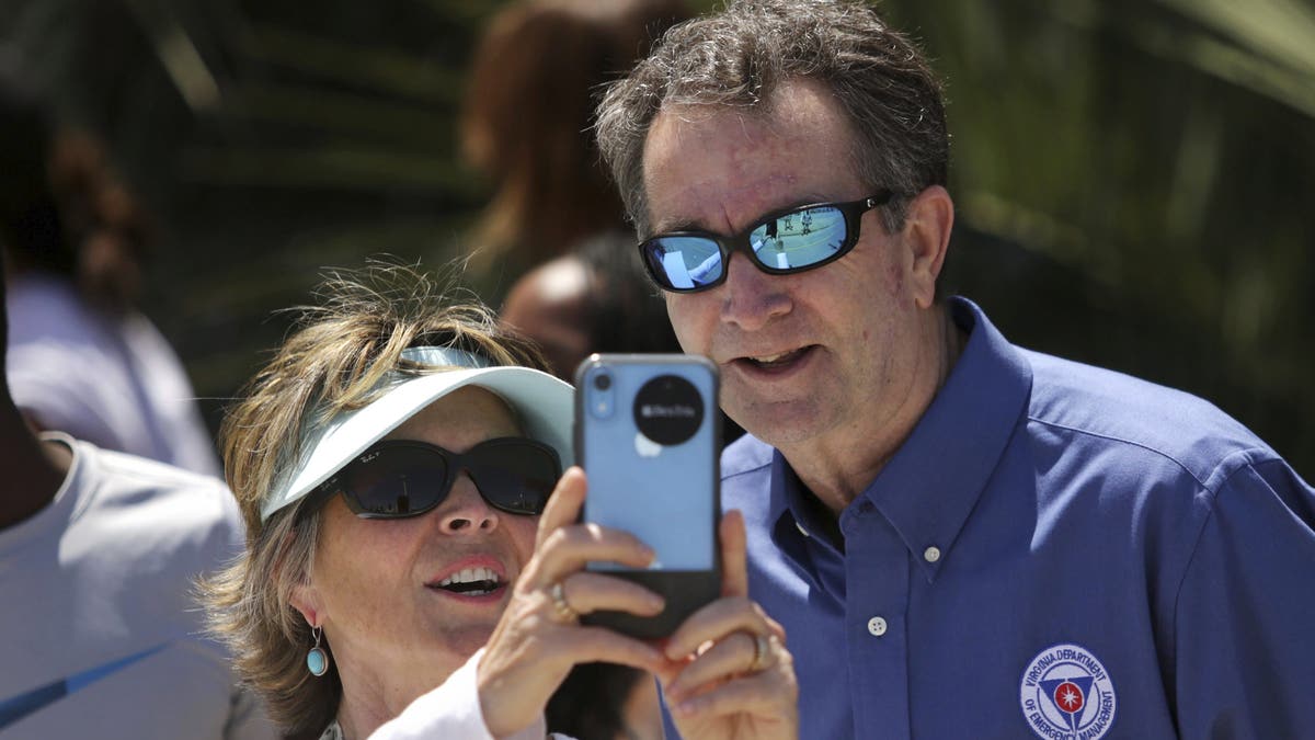 In this Saturday, May 23, 2020, photo, Gov. Ralph Northam and Tori Bloxom, of Onancock, Va., take a selfie as the governor visits the Oceanfront in Virginia Beach, Va., to see for himself how crowded the beach was. Northam has repeatedly urged Virginia residents to cover their faces in public during the COVID-19 pandemic, but the Democrat didn’t heed his own plea when he posed mask-less for photographs alongside residents during the weekend beach visit. A spokeswoman for the governor’s office said on Sunday that Northam should have brought a face mask with him during his visit on Saturday to the Virginia Beach Oceanfront. (Stephen M. Katz/The Virginian-Pilot via AP)