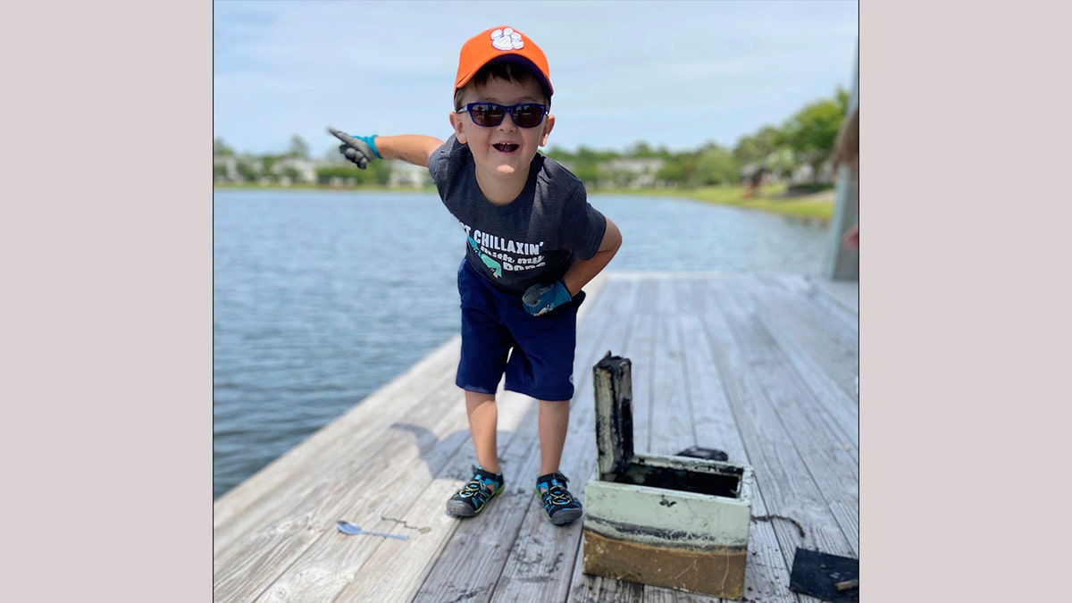 In this May 9, 2020, photo provided by Catherine Brewer, Knox Brewer stands next to a safe he pulled out of Whitney Lake in South Carolina. The 6-year-old was using a magnet attached to a string to fish for metal in the water when he reeled in a lockbox that police said was stolen from a woman who lived nearby eight years ago. (Catherine Brewer via AP)