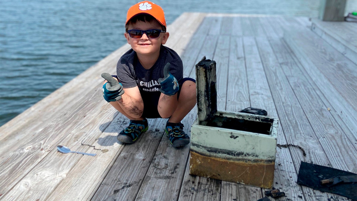 In this May 9, 2020, photo provided by Catherine Brewer, Knox Brewer poses next to a safe he pulled out of Whitney Lake in South Carolina. The 6-year-old was using a magnet attached to a string to fish for metal in the water when he reeled in a lockbox that police said was stolen from a woman who lived nearby eight years ago. (Catherine Brewer via AP)