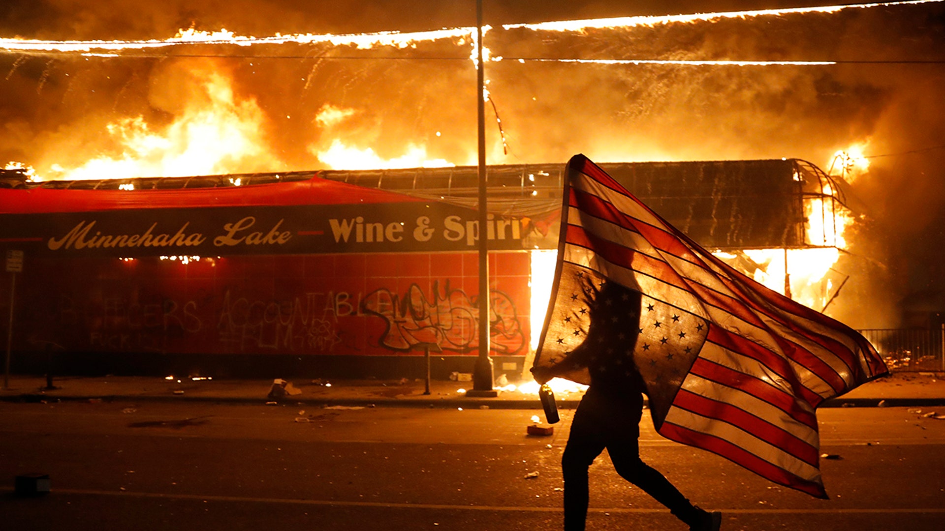 A protester carries a U.S. flag upside, a sign of distress, next to a burning building Thursday, May 28, 2020, in Minneapolis. Protests over the death of George Floyd, a black man who died in police custody Monday, broke out in Minneapolis for a third straight night.