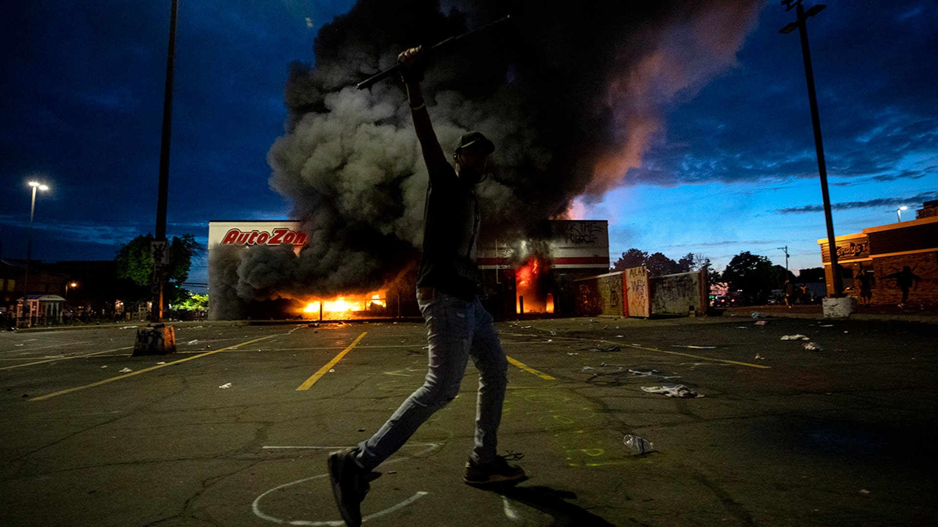 A man poses for a photo in the parking lot of an AutoZone store in flames, while protesters hold a rally for George Floyd in Minneapolis on Wednesday, May 27, 2020. Violent protests over the death of the black man in police custody broke out in Minneapolis for a second straight night Wednesday, with protesters in a standoff with officers outside a police precinct and looting of nearby stores.