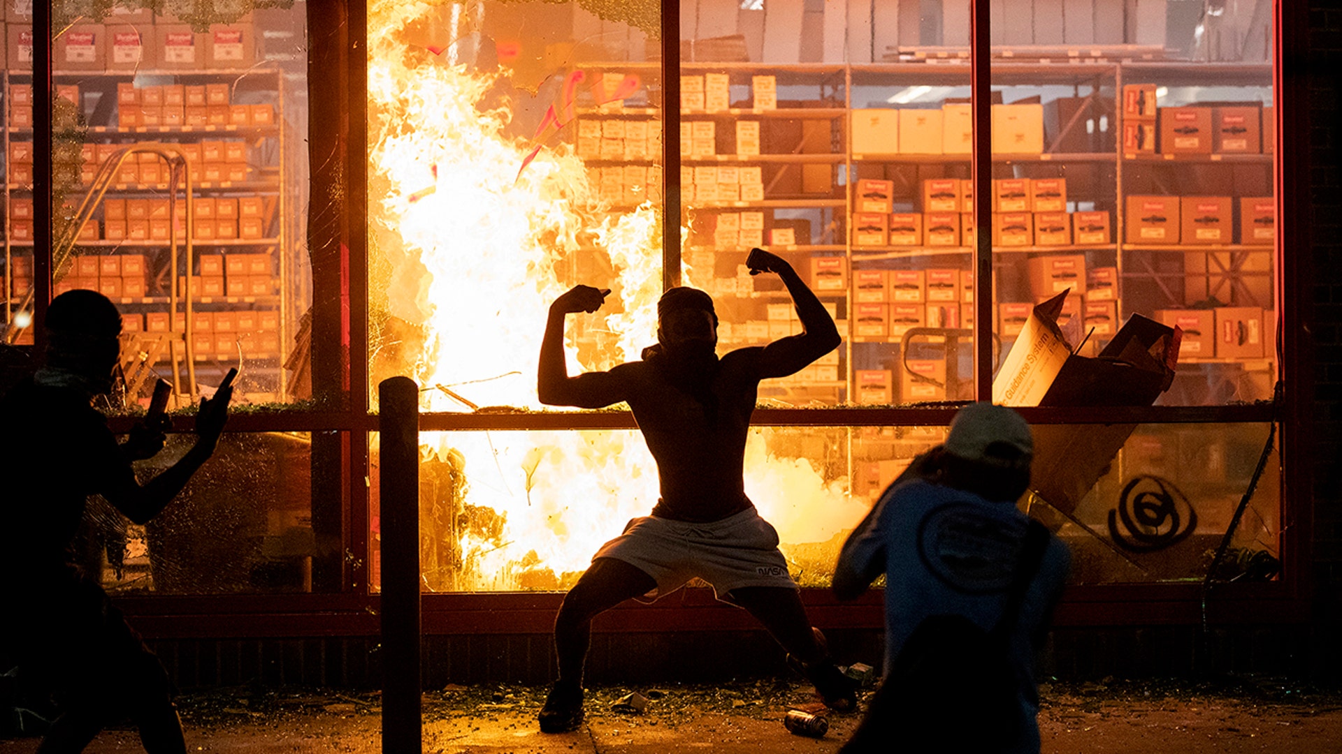 A man poses for photos in front of a fire at an AutoZone store, while protesters hold a rally for George Floyd in Minneapolis on Wednesday, May 27, 2020. 